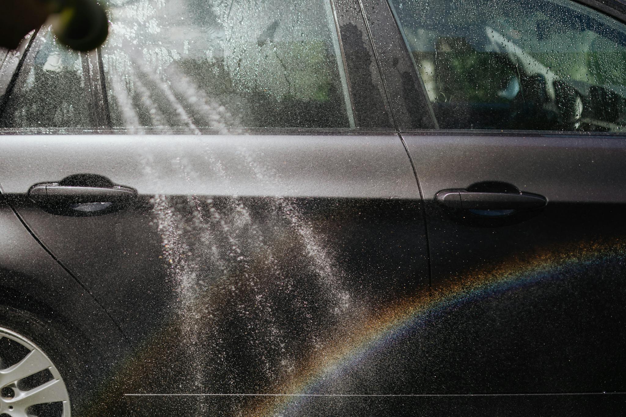 Close-up of a car being washed with a visible rainbow in sunlight.